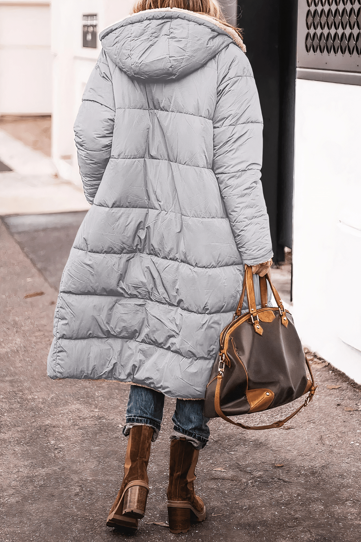 Person wearing a long light blue puffer coat with a hood, holding a brown leather handbag on a sidewalk.